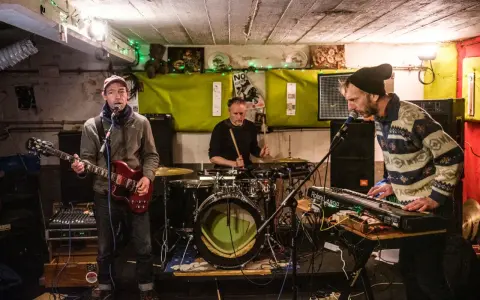 Un groupe de musique punk des années 90 joue dans un local sombre. Un guitariste, un batteur et un claviériste sont sur scène.