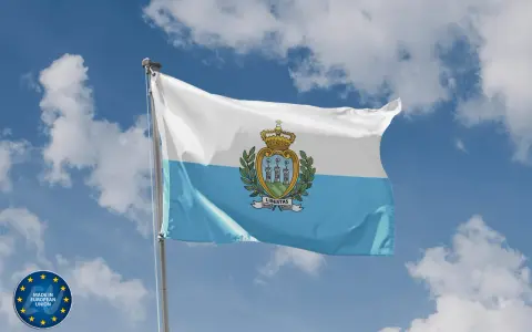 Flaga San Marino powiewa na tle błękitnego nieba z chmurami. Herb z trzema wieżami i koroną zdobi biało-błękitny materiał.