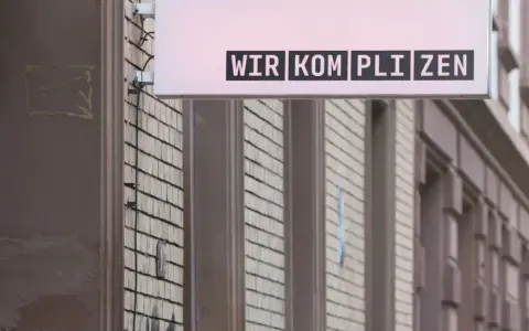 Leuchtreklame mit dem Schriftzug "WIR KOMPLIZEN" über einer Fassade.