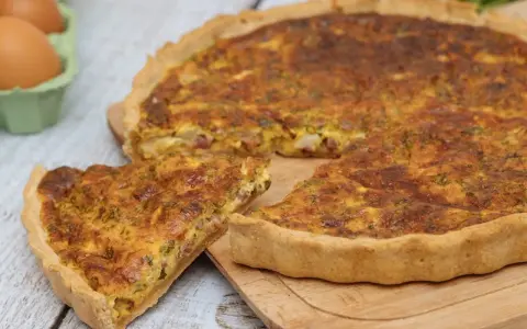 Une part de quiche lorraine, plat originaire de Lorraine, dorée et appétissante, sur une planche en bois. Des œufs frais sont visibles en arrière-plan.