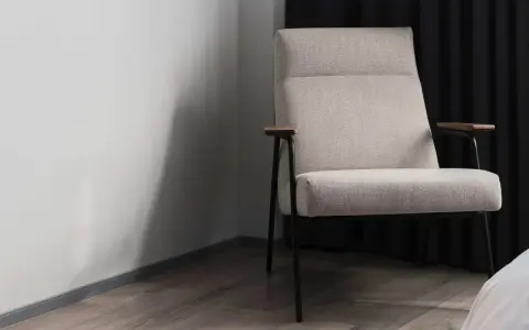 Fauteuil moderne avec un élégant tissu assise chaise, pieds fins et accoudoirs en bois. Il est placé près d'un mur blanc et de rideaux sombres.
