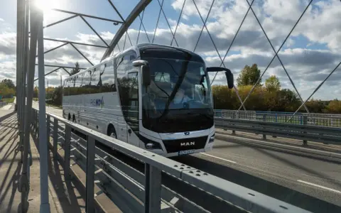 Biały autokar PKS NOVA jedzie przez most, pokonując trasę Warszawa-Augustów.