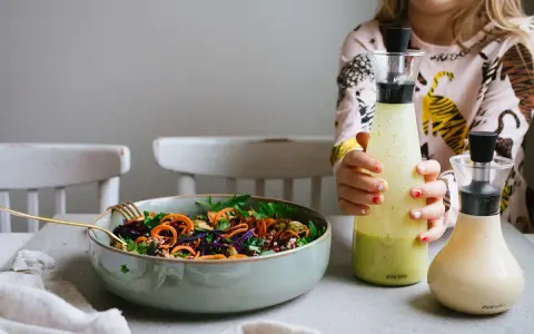 En skål med färgglad sallad och två flaskor dressing till sallad. En flicka håller i den ena flaskan.