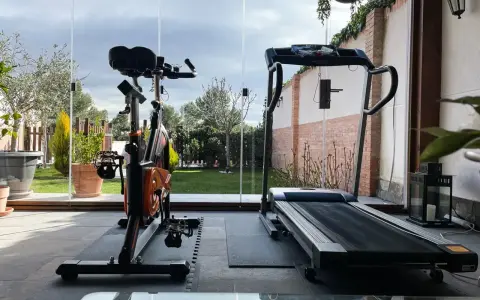 Un gimnasio en terraza con bicicleta estática y cinta de correr, con vistas a un jardín verde y un cielo nublado.