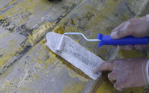 Händer med handskar arbetar med glasfibermatta och en roller för att laga en skadad yta.