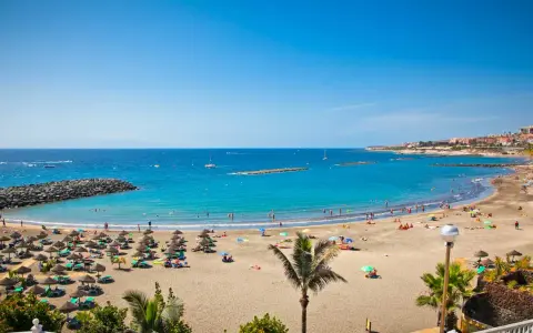Spiaggia affollata con ombrelloni e lettini, mare azzurro e città costiera sullo sfondo. Sembra l'isola più bella delle Canarie.