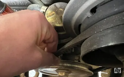 Hand prüft Keilriemen, der beim Anfahren quietscht. Der Riemen läuft über mehrere Riemenscheiben im Motorraum.