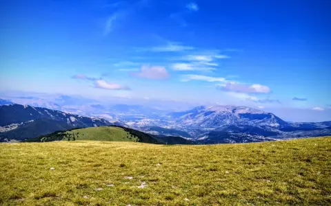 Prato verdeggiante con vista panoramica sul monte Pizzalto e le montagne circostanti sotto un cielo azzurro con nuvole sparse.