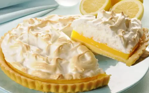 Puszysta tarta cytrynowa z bezą, z kawałkiem na łopatce. Obok leżą świeże cytryny.