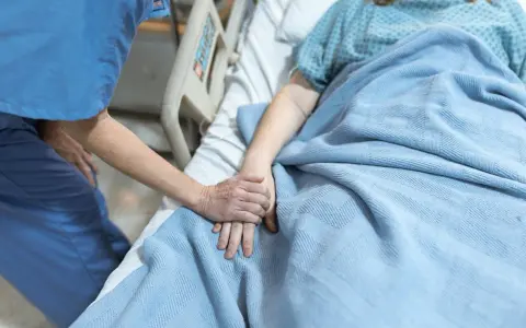 Pielęgniarka trzyma za rękę pacjentkę leżącą w łóżku szpitalnym pod niebieskim kocem.