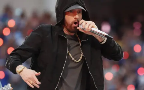 Eminem w czarnej bluzie z kapturem śpiewa do mikrofonu, w złotym łańcuchu i zegarku.
