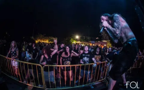 Un cantante con tatuajes canta en un concierto de charca rock. El público, animado, se agolpa tras una valla amarilla.