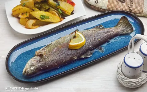 Eine ganze Forelle im Backofen, garniert mit einer Zitronenspalte, liegt auf einem blauen Teller. Daneben eine Beilage aus Kartoffeln und Gemüse.
