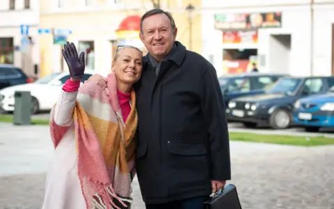 Małgorzata Szeptycka wita się z uśmiechem, obejmując partnera. W tle miejski krajobraz z samochodami.