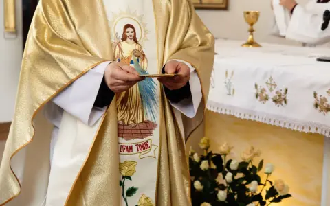 Kapłan w złotej szacie z wizerunkiem Jezusa trzyma hostię. Czy dzisiaj trzeba iść do kościoła?