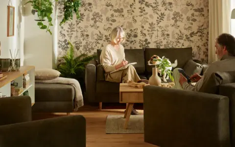 Przytulny salon, gdzie kobieta pisze, a mężczyzna korzysta z tabletu. Idealne inspiracje, jak urządzić pokój dla seniora, z wygodnymi meblami i roślinnością.