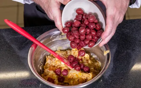 Hände geben Kirschen in eine Schüssel mit Teig für ein einfaches Kirschenmichel Rezept. Ein roter Teigschaber liegt daneben.