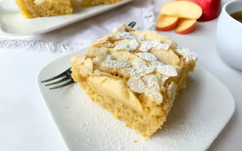 Ein Stück saftiger Apfelkuchen mit Apfelmus, bestreut mit Puderzucker und Mandelblättchen, wartet auf dem Teller.