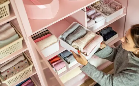 Frau sortiert Kleidung in einem rosafarbenen Schrank. Die Fächer sind ordentlich mit gefalteten Kleidungsstücken gefüllt, die nach Farben sortiert sind, um eine harmonische Reihenfolge zu schaffen.
