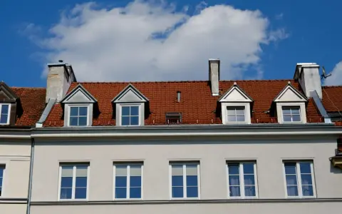Czerwony dach z lukarnami i kominami nad jasnoszarą elewacją.