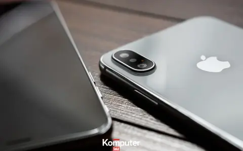 Dwa telefony leżą obok siebie na drewnianym blacie. Jeden z nich, srebrny iPhone, ma widoczny moduł aparatu. Drugi, z ciemnym ekranem, jest ustawiony bokiem. Idealne do rozmowy przez **Facetime**.