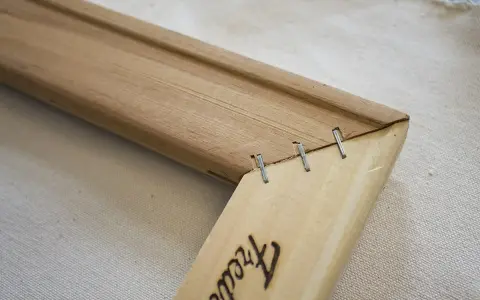 Détail d'un coin de cadre en bois, montrant comment tendre une toile sans châssis avec des agrafes.
