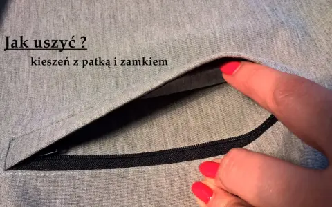Szara tkanina z widocznym zamkiem i patką. Dłoń z czerwonym lakierem do paznokci wsuwa się do kieszeni z patką.