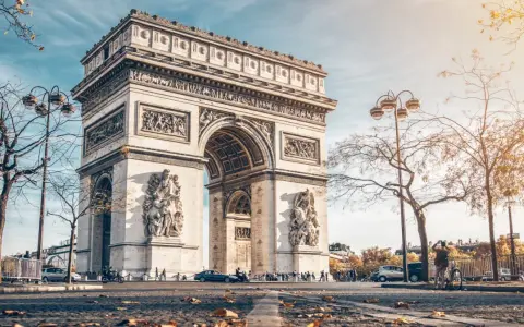 La majestuosa entrada del Arco del Triunfo en París, con esculturas y detalles ornamentales, bajo un cielo azul y árboles otoñales.