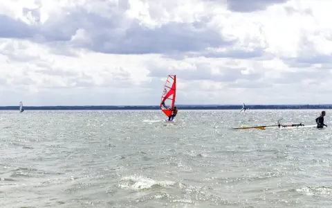 Czerwony żagiel do windsurfingu na wodzie, surfer w akcji. W tle inni windsurferzy i chmury.