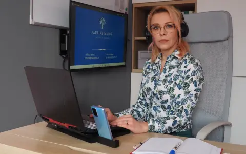 Kobieta w okularach i słuchawkach siedzi przy biurku z laptopem, telefonem i notatnikiem.