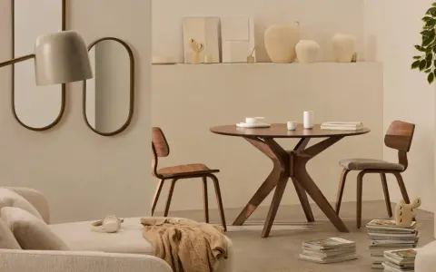 Dos espejos ovalados cuelgan sobre un sofá beige. A la derecha, una mesa redonda de madera con sillas y un jarrón.