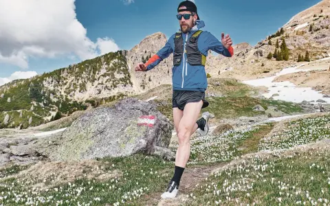 Uomo con barba corre in montagna, circondato da crochi bianchi e rocce. Il sentiero si snoda tra prati e pendii innevati.
