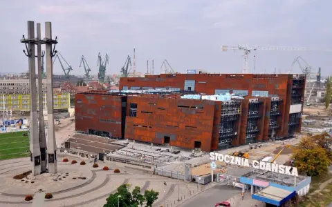 Nowoczesne muzeum na terenie Stoczni Gdańskiej, z charakterystycznymi, rdzawymi fasadami, w otoczeniu dźwigów stoczniowych i Pomnika Poległych Stoczniowców.