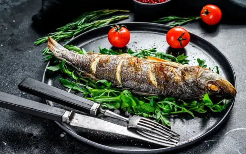 Un bar entier, parfaitement doré, est prêt pour la dégustation. Ce plat de cuisson bar au four est servi sur un lit de roquette fraîche, accompagné de tomates cerises et de romarin.
