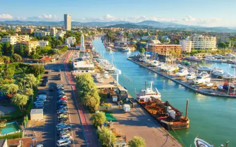 Vista aerea del porto di Rimini con barche ormeggiate, edifici e montagne sullo sfondo.