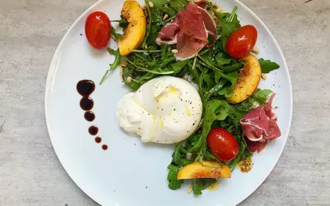 Burrata mit Rucola, Tomaten, Pfirsichspalten und Prosciutto auf einem Teller.