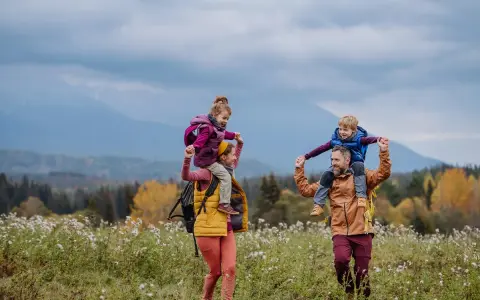 Rodzina na spacerze po łące. Dzieci siedzą na ramionach rodziców, jakby hasające zające zakopane w trawie.