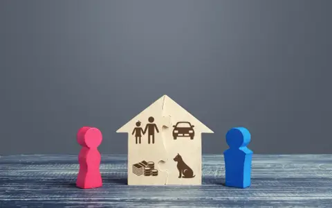 Drewniany dom z puzzli z symbolami rodziny, pieniędzy, samochodu i psa, między dwiema figurkami.