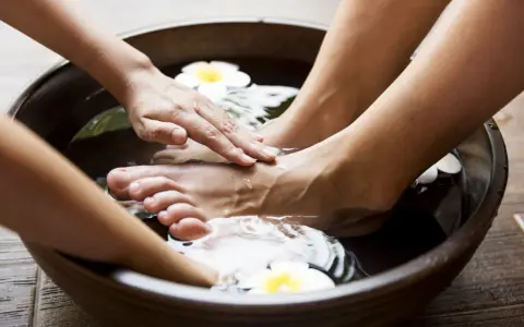 Soaking feet in a warm bath with Epsom salt for feet infection. A hand gently massages the foot, surrounded by water and white flowers.
