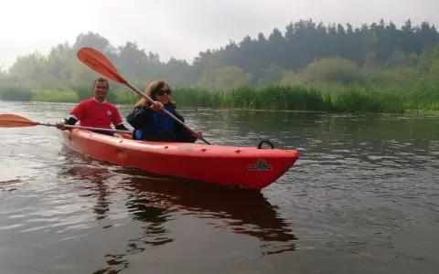 Para płynie kajakiem po rzece, w tle las i trzciny. To piękny dzień na spływ Pilicą, miasto w oddali.