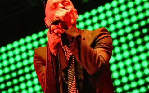 Wokalista REM, Michael Stipe, śpiewa na scenie, gdy światła migoczą na zielono.