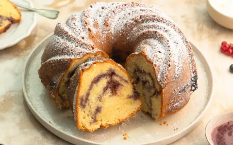 Ein Gugelhupf mit Marmelade, bestäubt mit Puderzucker. Ein Stück ist bereits angeschnitten und zeigt die köstliche Füllung.