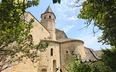 Vue d'une église romane à Villeneuve de Rouergue, encadrée par des arbres verdoyants sous un ciel bleu parsemé de nuages.