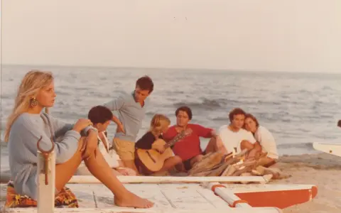 Giovani su una spiaggia, un gruppo di amici che ricorda i film estivi italiani, con chitarra e falò.