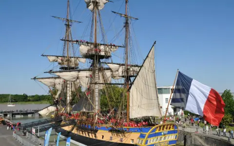 Le majestueux bateau l'Hermione, réplique d'une frégate du XVIIIe siècle, est amarré, pavoisé du drapeau français. Une foule s'est rassemblée pour admirer ce navire historique.