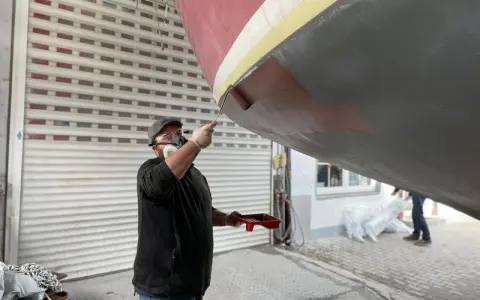 Mann mit Maske und Schutzbrille streicht mit einer Rolle den Rumpf eines Bootes.