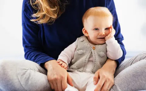 Uśmiechnięte dziecko w sweterku, które zaczyna siadać, siedzi na kolanach mamy.