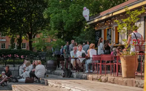 Folk njuter av solen på uteserveringar i Stockholm. En glasskylt syns ovanför en servering.