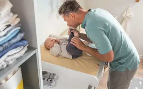 Tata całuje nóżki dziecka na przewijaku. W tle stos ubranek i kosz. Jaki przewijak wybrać?