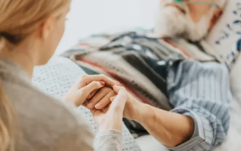 Mani che si stringono tra una giovane donna e un anziano a letto, con cannule nasali. Il caregiver viene pagato per la sua dedizione.